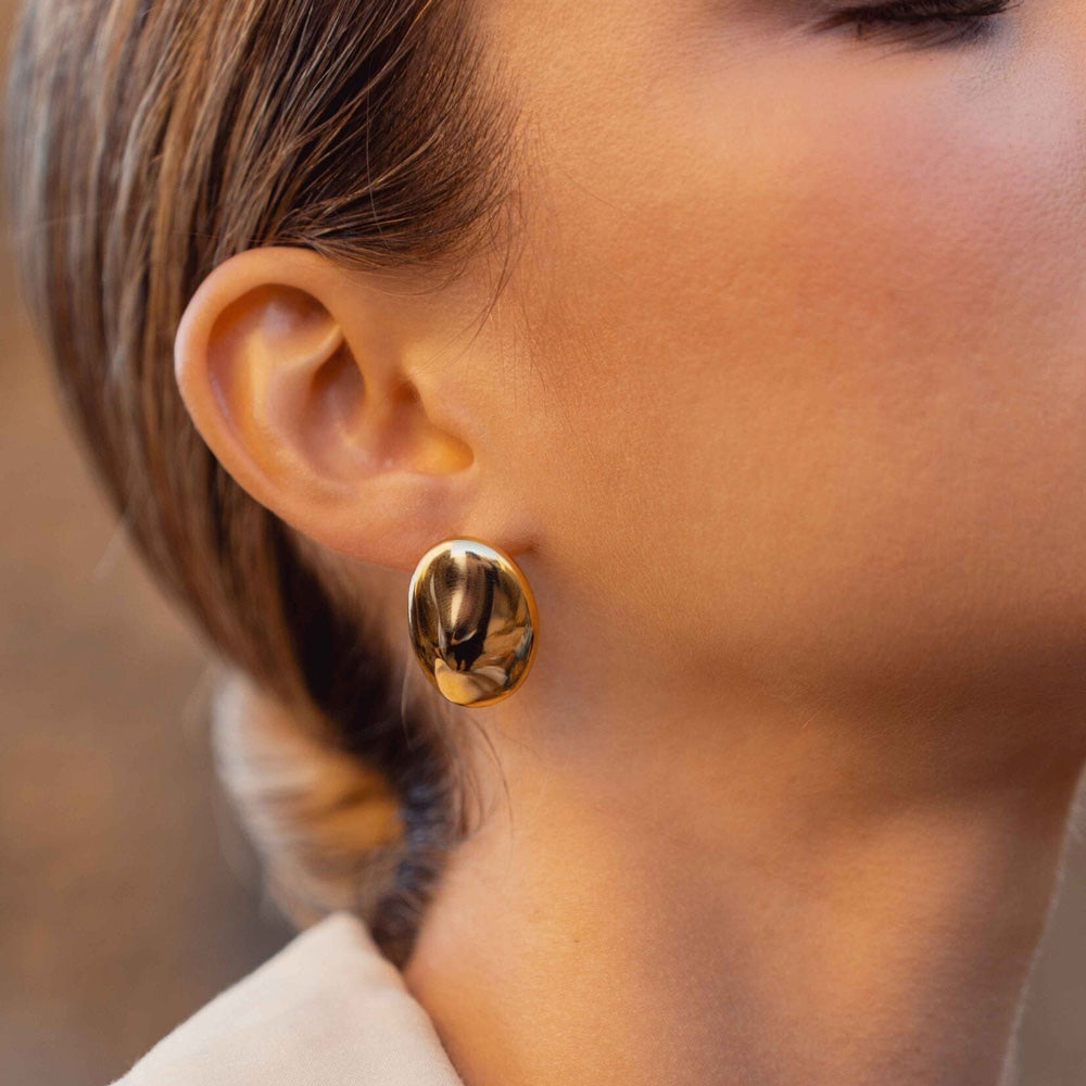 Kullanväriset suuret ovaalit nappikorvakorut peilipinnalla korvassa.