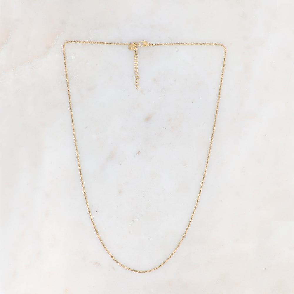 Siro pitkä kaulakoru kullanvärisenä vaalealla pohjalla.