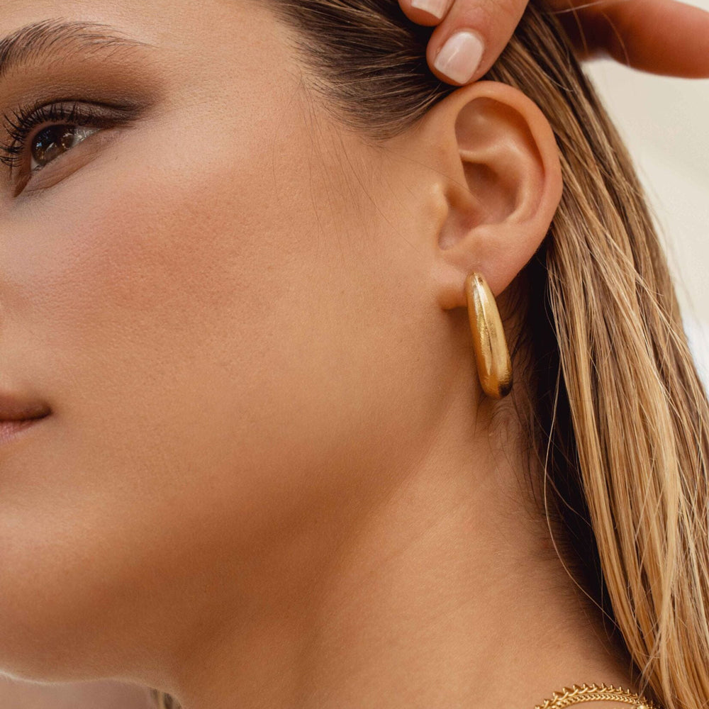 Modernit kultaiset ovaalit rengaskorvakorut harjatulla pinnalla mallin korvassa.