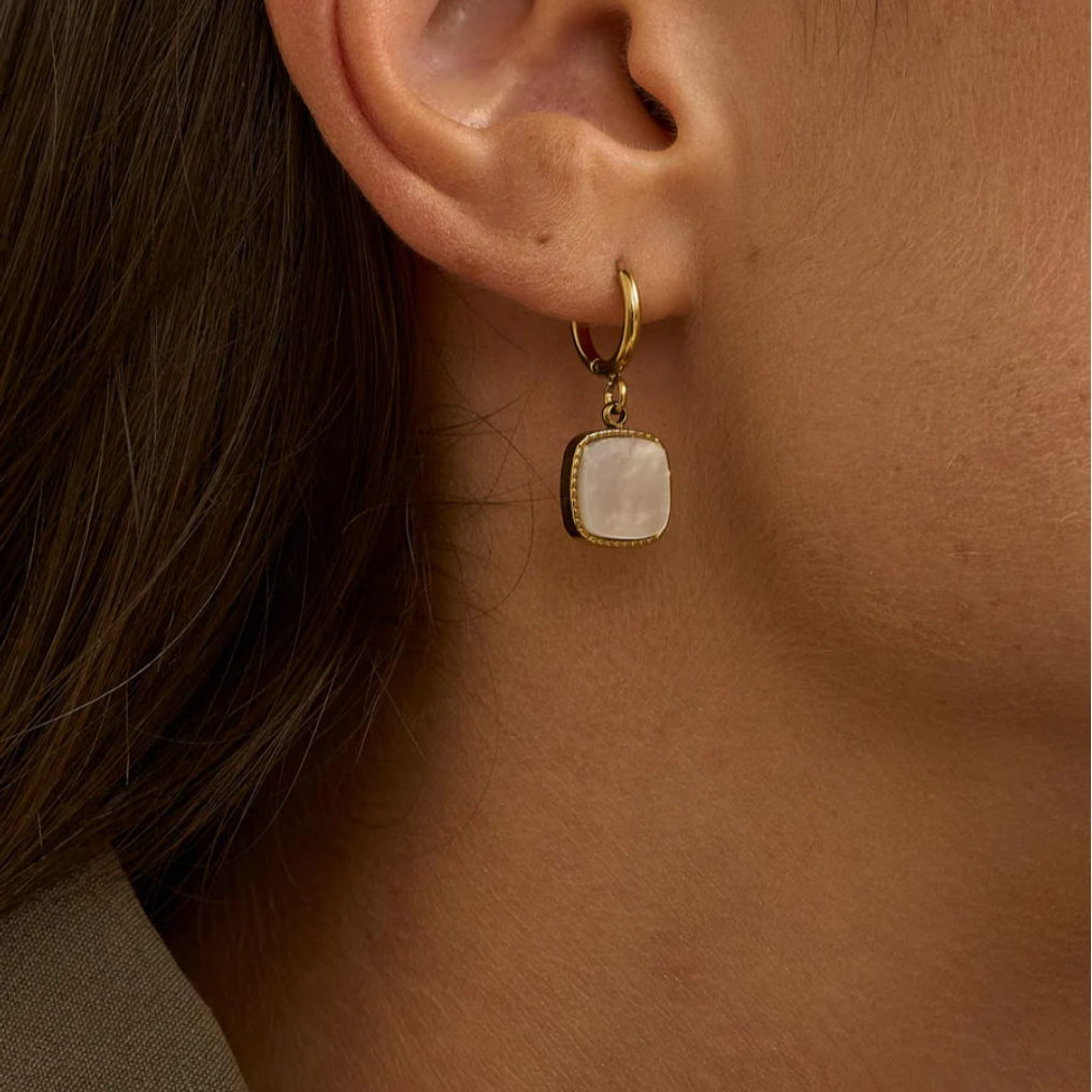 Kultaiset rengaskorvakorut valkoisella helmiäisriipuksella korvassa.